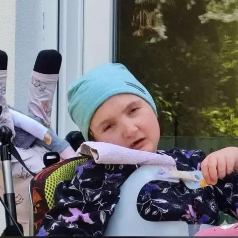 Kind mit türkisfarbener Mütze sitzt in einem Therapiestuhl am Fenster